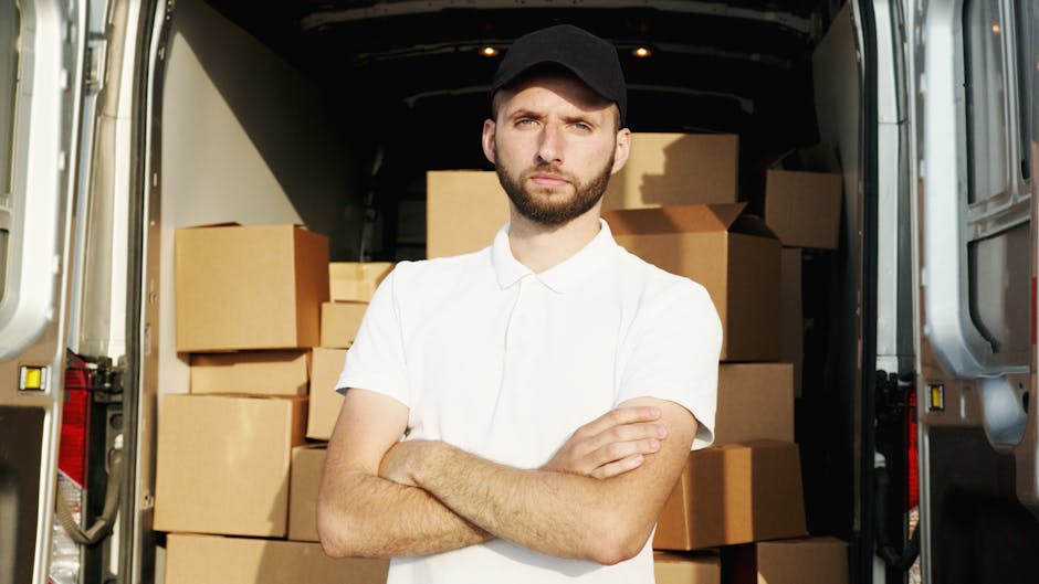 A young man dressed in a white polo shirt stands inside the rear of a moving van with his arms crossed, surrounded by stacked cardboard boxes of various sizes wrapped in protective material. The boxes are arranged closely together, occupying the entire interior space of the van, which is lit by overhead lighting. The man has a serious expression, with a beard and a black cap, as he appears to oversee the home relocation process. Outside the van, the environment suggests a loading area or driveway. This scene illustrates furniture transport and packing during a professional removals service, with equipment such as straps and blankets potentially used for securing items during loading and unloading. Man with Van Harrow, a company specializing in removals, is involved in the logistics of packing and moving belongings efficiently and securely.