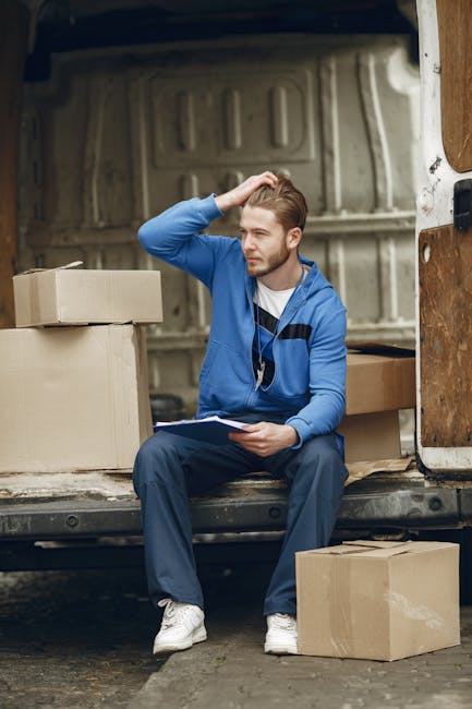 A young man with light brown hair, wearing a blue jacket, dark trousers, and white sneakers, is sitting on the edge of an open moving van with a beige interior, parked outdoors on a paved surface. He is holding a clipboard in his lap and appears to be contemplating or reviewing a list, with his right hand resting on his head. Around him are several cardboard boxes, some sealed and others partially open, positioned both inside the van and on the ground nearby. Inside the van, visible behind him, are more packed cardboard boxes, some with packing tape, alongside a few wrapped items covered with plastic film or blankets. The scene illustrates a home relocation process in progress, emphasizing the careful planning needed during furniture transport and packing, which are key aspects of professional removals services like those offered by Man with Van Harrow.