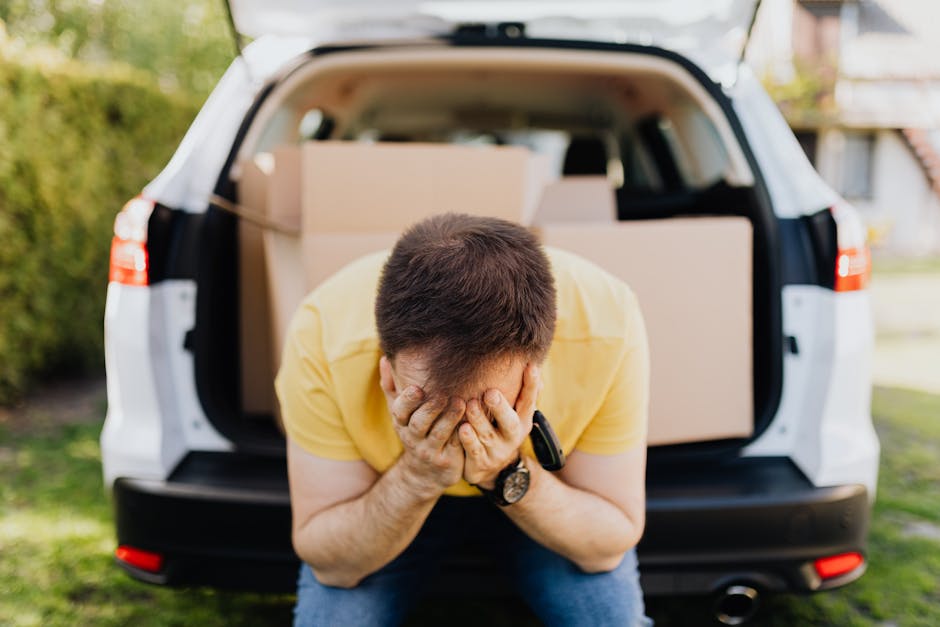 A young man with light brown hair, wearing a blue jacket, dark trousers, and white sneakers, is sitting on the edge of an open moving van with a beige interior, parked outdoors on a paved surface. He is holding a clipboard in his lap and appears to be contemplating or reviewing a list, with his right hand resting on his head. Around him are several cardboard boxes, some sealed and others partially open, positioned both inside the van and on the ground nearby. Inside the van, visible behind him, are more packed cardboard boxes, some with packing tape, alongside a few wrapped items covered with plastic film or blankets. The scene illustrates a home relocation process in progress, emphasizing the careful planning needed during furniture transport and packing, which are key aspects of professional removals services like those offered by Man with Van Harrow.