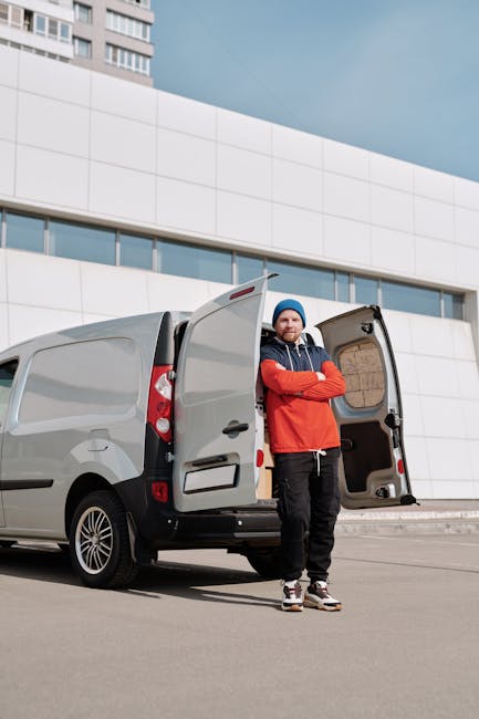 A man dressed in a red and black jacket, wearing a blue beanie, stands arms crossed beside a grey delivery van with its rear doors open, revealing an empty interior. The van is parked on a paved area in front of a modern white building with curved architectural features and large windows. The scene takes place outdoors during daytime with a partly cloudy sky. The man appears to be preparing for or concluding a home relocation or furniture transport task, which is a typical service offered by Man with Van Harrow, as referenced on the webpage about parking suspensions for removals. The image highlights transportation and logistical aspects of house removals, including loading or unloading equipment, with the van positioned close to the property for efficient moving process.