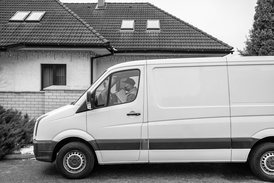 A white panel van parked on the street in front of a residential house with a pitched roof, dormer windows, and string lights hanging from the eaves. Inside the van, a person wearing a face mask is seated in the driver's seat, reading a document. The van's side door is closed, and no loading or unloading actions are visible. The house has a brick wall surrounding a small garden area, with shrubbery visible on the side. The scene is captured during daytime in a suburban neighbourhood, illustrating a typical home relocation or moving process, with the van ready for furniture transport and packing as part of a house removals service, consistent with the offerings of Man with Van Harrow.