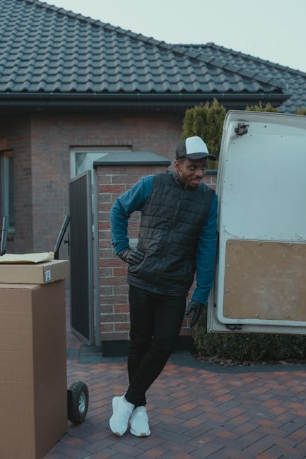 A man wearing a blue long-sleeved shirt, a dark padded vest, and a baseball cap stands beside an open van door during a home relocation process. He has gloves on and is leaning slightly with his hands on his hips, looking at the loading area. The van, positioned on a paved driveway in front of a brick house with a tiled roof, is being used by Man with Van Harrow for furniture transport and packing and moving services. Nearby, there is a large cardboard box on a trolley and some packing materials, suggesting ongoing packing or loading activities. The environment appears well-lit, likely early evening or late afternoon, with greenery visible behind the house. This scene captures the logistics involved in professional house removals within the Harrow area, highlighting the careful handling and organization typical of Man with Van Harrow's service process.