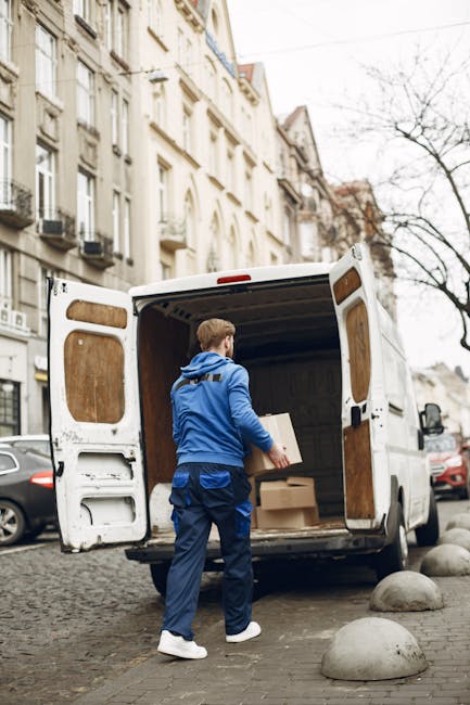A man wearing a blue jacket and dark trousers is loading cardboard boxes into the rear of a white delivery van parked on a cobblestone street in North Harrow. The van's rear doors are open, revealing an empty interior with wooden paneling and some packing materials. The street is lined with multi-storey residential buildings with light-colored facades, balconies, and large windows, typical of urban house removals. Several parked cars are visible along the curb, and leafless trees indicate a cooler season. The scene captures the process of home relocation, with the man carefully placing boxes into the van for transportation and moving services offered by Man with Van Harrow. Concrete bollards are positioned along the edge of the sidewalk to prevent vehicle overreach, and natural daylight illuminates the scene, emphasizing the practical aspects of furniture transport and packing during a house move.