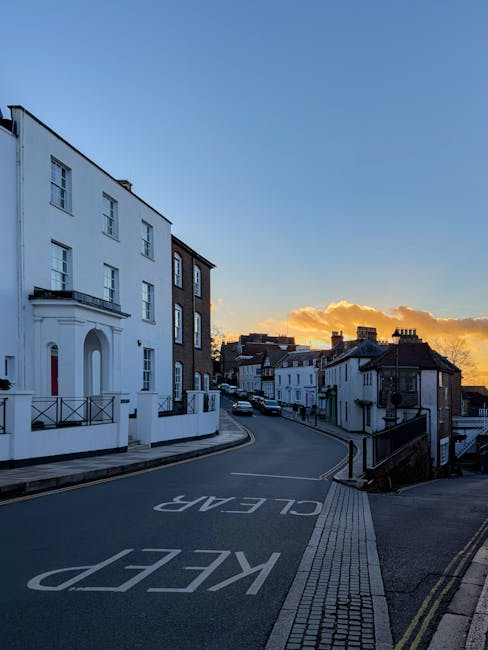 A narrow residential street in North Harrow during sunset, with the sky displaying a gradient of blue and orange clouds. On the left side, there is a large white building with multiple windows, a small balcony, and a red door, indicative of typical house removal locations. The street curves gently upward, with parked cars visible further along the road. The pavement is lined with a tactile paving strip and double yellow lines along the curb, with a clear 'KEEP CLEAR' marking painted on the road surface close to the foreground. This setting reflects a quiet area suitable for house removals and furniture transport services, often requiring careful loading and unloading processes near residential properties. The overall scene emphasizes the calm environment suitable for operational logistics by professional movers like Man with Van Harrow.