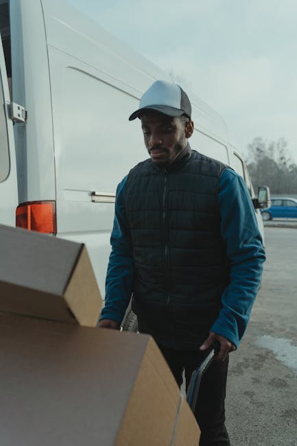 A man wearing a light grey cap, a navy blue padded jacket over a blue long-sleeve shirt, is standing outdoors on a cloudy day, next to a white moving van. He is holding a small hand truck or dolly, which is partially visible, and is positioned near several brown cardboard boxes of various sizes, some stacked on top of each other. The loading area of the van is open, showing the interior, and the ground is made of asphalt. In the background, there are a few other vehicles, including a blue car, and trees with sparse foliage, indicating a typical house removal scene. This image illustrates the process of packing and preparing for a home relocation, with careful handling of packed boxes, and is associated with professional removals services provided by Man with Van Harrow, focusing on furniture transport and moving logistics.
