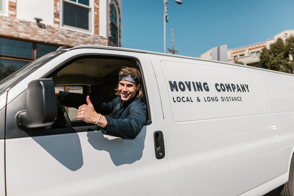 A smiling young man with long hair, wearing a patterned headband and dark clothing, is sitting in the driver's seat of a white moving van with the words 'MOVING COMPANY LOCAL & LONG DISTANCE' printed on the side. He is giving a thumbs-up gesture through the open window, indicating a positive attitude towards the home relocation and furniture transport process. The van is parked outside a modern building with large windows and brick facades, under a clear blue sky. The image captures the loading or unloading phase of a house removal, with the vehicle positioned on a paved street suitable for logistics and moving services by Man with Van Harrow. The scene emphasizes the professional approach to packing and moving, highlighting the vehicle used for transport and the readiness of the team involved in the house removals or office relocations.