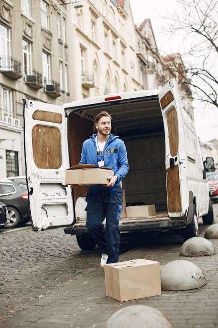 A man wearing a light grey cap, a navy blue padded jacket over a blue long-sleeve shirt, is standing outdoors on a cloudy day, next to a white moving van. He is holding a small hand truck or dolly, which is partially visible, and is positioned near several brown cardboard boxes of various sizes, some stacked on top of each other. The loading area of the van is open, showing the interior, and the ground is made of asphalt. In the background, there are a few other vehicles, including a blue car, and trees with sparse foliage, indicating a typical house removal scene. This image illustrates the process of packing and preparing for a home relocation, with careful handling of packed boxes, and is associated with professional removals services provided by Man with Van Harrow, focusing on furniture transport and moving logistics.