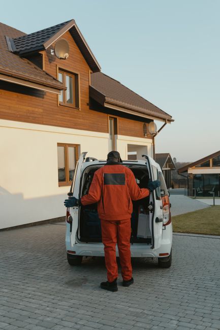 A mover dressed in a red work uniform with black accents is standing at the rear of a small white van, loading or unloading household items. The van is parked on a paved driveway in front of a modern,