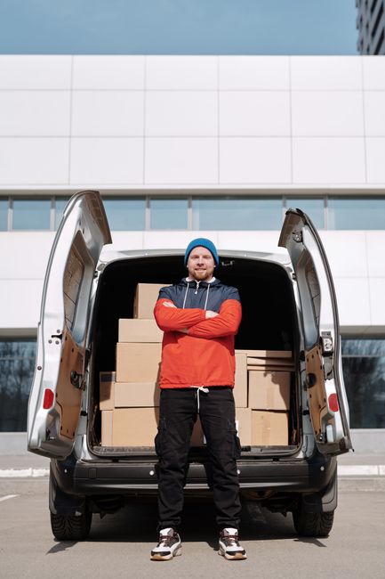 A man wearing a blue beanie, red and navy jacket, and black trousers stands with arms crossed in front of an open van trunk filled with cardboard boxes during a home relocation. The boxes are stacked neatly inside the van, which is parked on a paved surface in front of a modern building with large glass windows and a white facade. The scene depicts the loading process in progress, with the man possibly overseeing the furniture transport or packing and moving operations. The van's rear doors are fully open, revealing the interior filled with packed items prepared for moving. This image illustrates the logistics of a house removals service, highlighting the use of protective packaging materials and large vehicles essential for efficient and careful furniture transport, as provided by Man with Van Harrow.