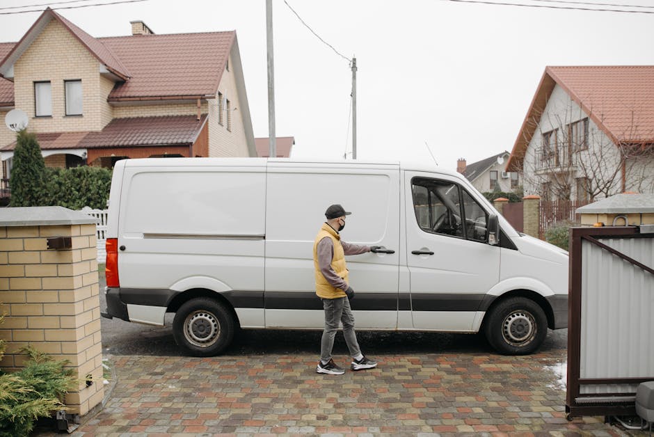 A man wearing a black cap, glasses, a yellow and gray jacket, and dark trousers stands on a cobbled driveway, opening the sliding side door of a white commercial van for a home relocation or furniture transport service. The van is parked on the street in front of a residential property with a brick wall and a gate to the right. In the background, there are two detached houses with pitched roofs covered in red tiles, one with beige brickwork and the other with white exterior walls, surrounded by green hedges and trees. The scene appears to be overcast with diffused natural light, and the overall environment suggests the loading process during a house removal, with visible elements such as cardboard packing materials and a clear focus on the vehicle's interior access, supporting efficient packing and moving operations as provided by Man with Van Harrow.