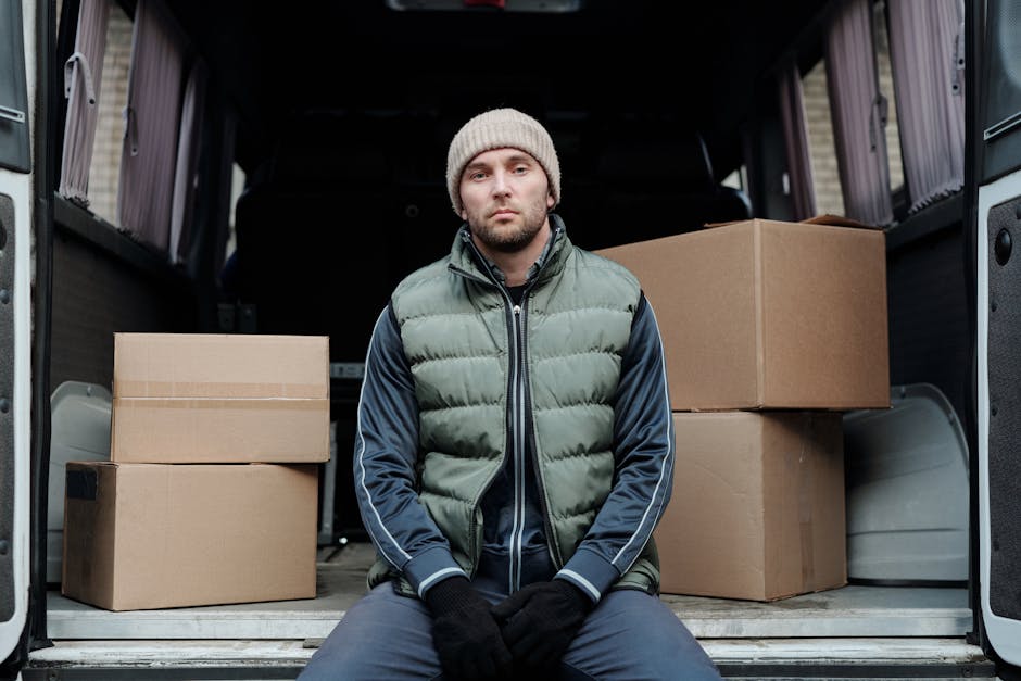 A man wearing a blue beanie, red and navy jacket, and black trousers stands with arms crossed in front of an open van trunk filled with cardboard boxes during a home relocation. The boxes are stacked neatly inside the van, which is parked on a paved surface in front of a modern building with large glass windows and a white facade. The scene depicts the loading process in progress, with the man possibly overseeing the furniture transport or packing and moving operations. The van's rear doors are fully open, revealing the interior filled with packed items prepared for moving. This image illustrates the logistics of a house removals service, highlighting the use of protective packaging materials and large vehicles essential for efficient and careful furniture transport, as provided by Man with Van Harrow.