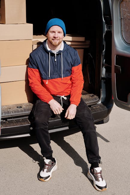 A young man with a beard, wearing a blue beanie hat and a navy and red jacket, is sitting on the edge of an open van trunk in a driveway. Inside the vehicle, there are multiple brown cardboard boxes stacked neatly, some covered with protective plastic wrap. The boxes vary in size and are positioned along the side of the van, which is parked close to a building. The man is wearing black trousers and white sneakers with black and red accents. The loading area outside the van appears to be a paved surface with shadows indicating daylight. The scene captures a moment during a home relocation process, with the man possibly preparing for or taking a break from packing and furniture transport tasks. Man with Van Harrow, a professional removals service, is involved in this furniture transport and packing process, supporting efficient house moves in the Harrow area.