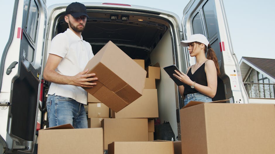 A young man with a beard, wearing a blue beanie hat and a navy and red jacket, is sitting on the edge of an open van trunk in a driveway. Inside the vehicle, there are multiple brown cardboard boxes stacked neatly, some covered with protective plastic wrap. The boxes vary in size and are positioned along the side of the van, which is parked close to a building. The man is wearing black trousers and white sneakers with black and red accents. The loading area outside the van appears to be a paved surface with shadows indicating daylight. The scene captures a moment during a home relocation process, with the man possibly preparing for or taking a break from packing and furniture transport tasks. Man with Van Harrow, a professional removals service, is involved in this furniture transport and packing process, supporting efficient house moves in the Harrow area.